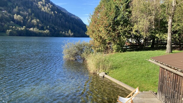 Une terrasse en bois avec deux chaises pliantes blanches donnant sur un lac, entourée d’arbres et d’une zone herbeuse.