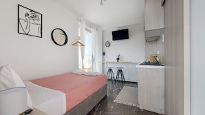 Et lille hotelværelse med en enkelt seng, et lille skrivebord, et fjernsyn og et tekøkken.