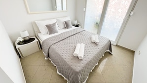 A neatly made bed with a grey quilt and pillows, a bedside table with a lamp, and a large window with a floral pattern.