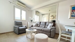 A living room with a grey sofa, a grey armchair, a white coffee table, and a white ottoman. There is a white side table with a vase of flowers. The room has large windows, an air conditioning unit, and a white wall with a painting.