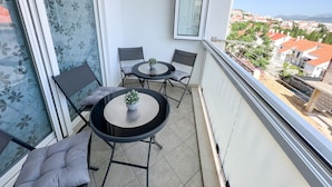 A balcony with two round tables, each with a chair, and a potted plant. The balcony has a view of a residential area with red-roofed houses.