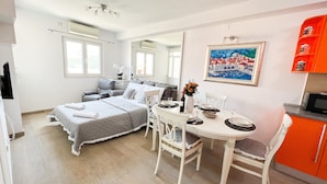 A compact bedroom with a bed, bedside table, and a small window. Adjacent is a dining area with a table, chairs, and a painting on the wall.