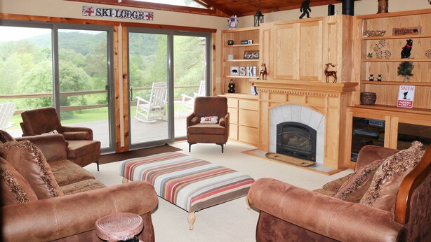 A cozy lodge interior with leather sofas, a fireplace, wooden shelves with ski-themed decor, and a large sliding glass door offering a scenic view. - Welcome to Howe Hill Retreat! (Great Valley)