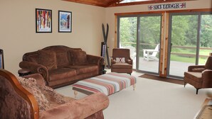 A cozy living room with a brown leather sofa, a striped ottoman, and a brown armchair. There are framed pictures on the wall, a ski lodge sign, and a large window offering a view of the outdoors. - Welcome to Howe Hill Retreat! (Great Valley)
