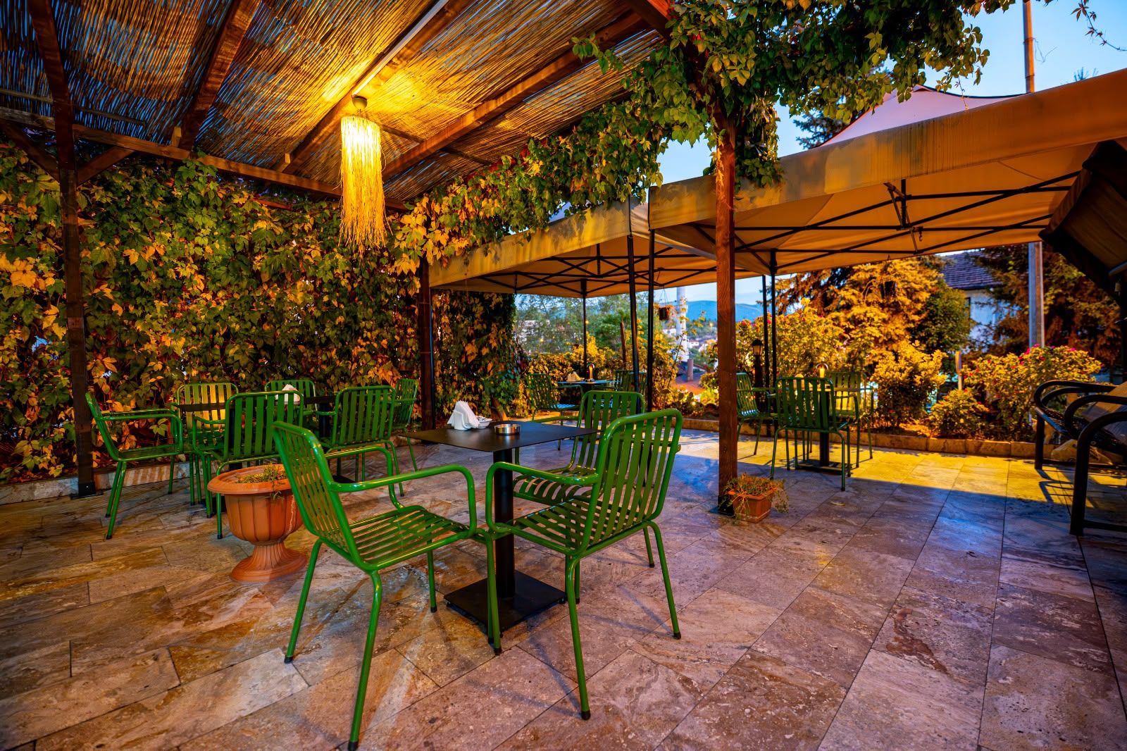 An outdoor seating area with green chairs and a table, surrounded by foliage and a pergola structure.