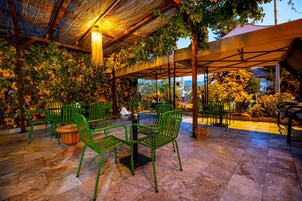 Un espacio al aire libre con sillas verdes y una mesa, rodeado de follaje y una estructura de pérgola.