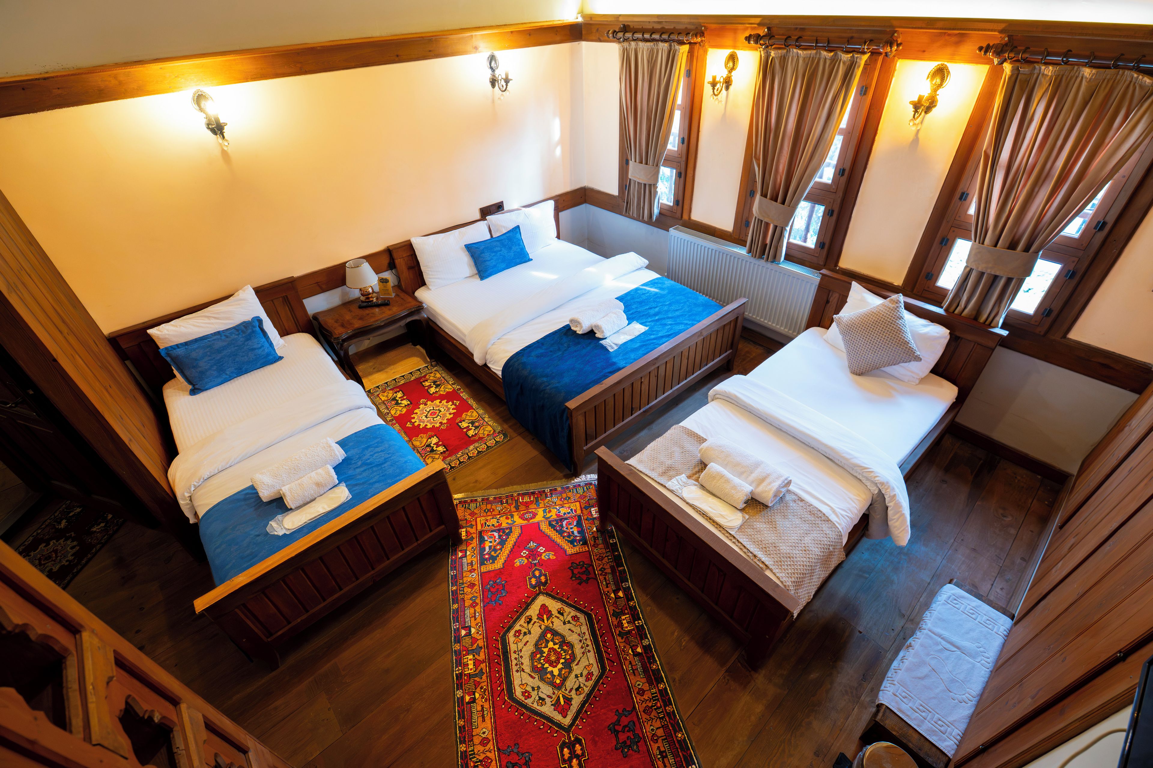 A hotel room with three beds, a wooden floor, a red patterned rug, and a window with curtains.