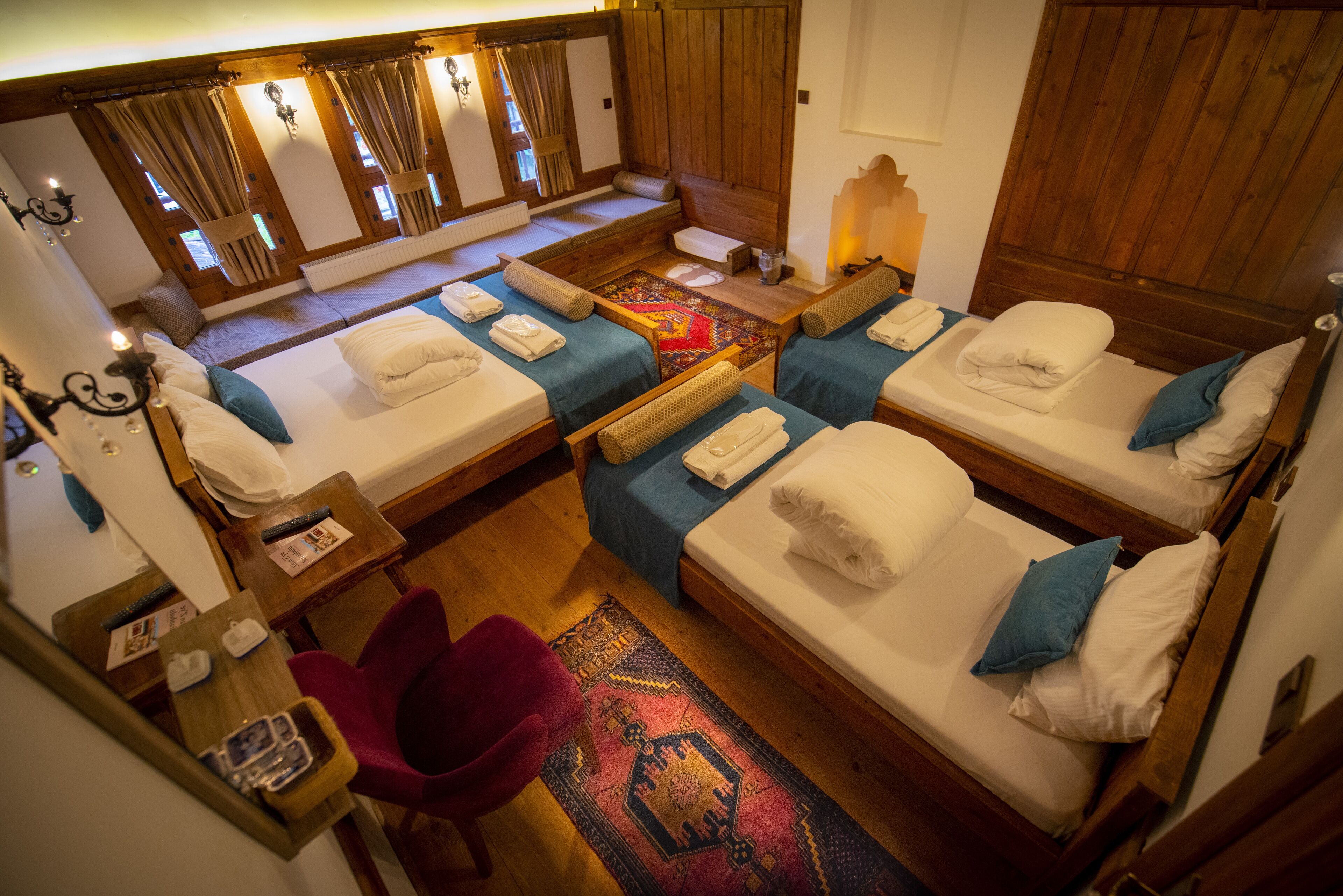 A room with four beds arranged in a U-shape, each with a pillow and a folded towel. There is a small table with a lamp and a chair.