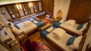 A room with four beds arranged in a U-shape, each with a pillow and a folded towel. There is a small table with a lamp and a chair.