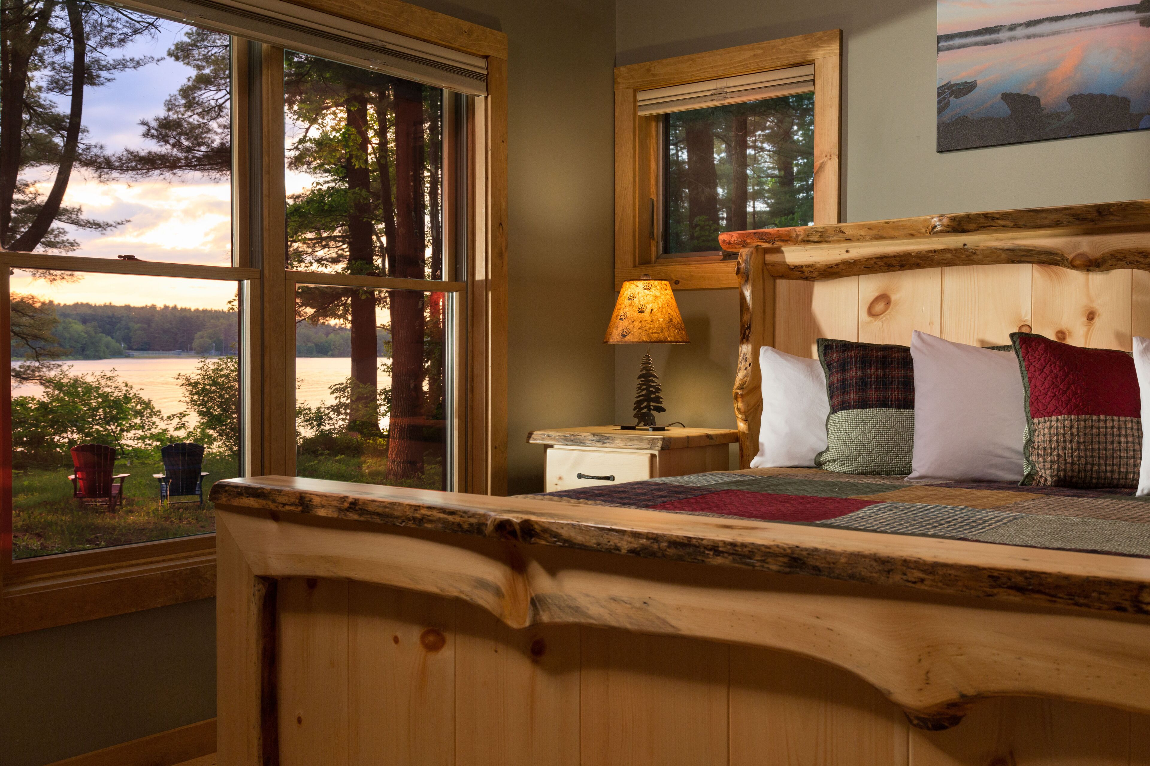 Ein Bett mit Holzkopfteil, ein Nachttisch mit Lampe und Blick auf einen See durch große Fenster.