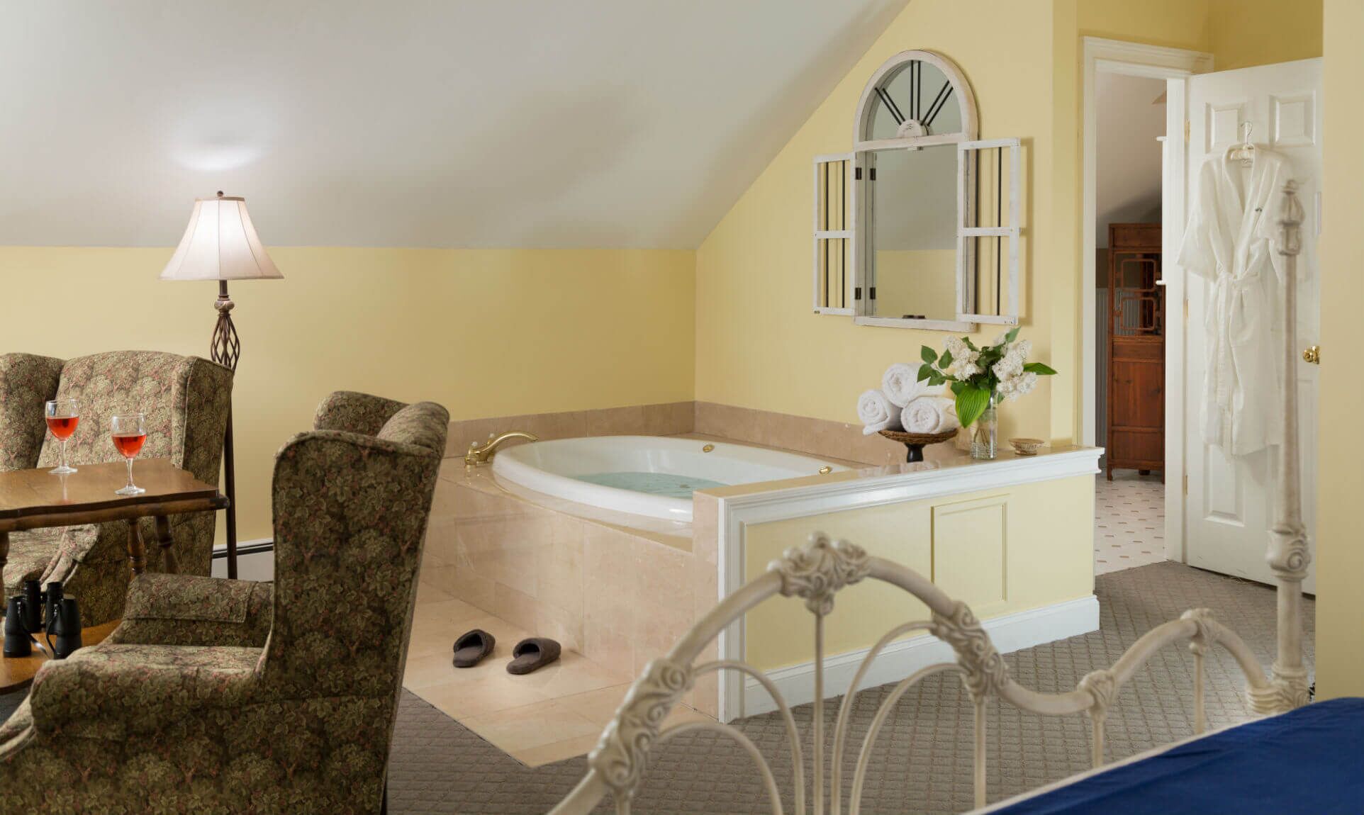 A bedroom with a large bathtub, a vanity with a mirror, a bed, and a table with two glasses of red wine.