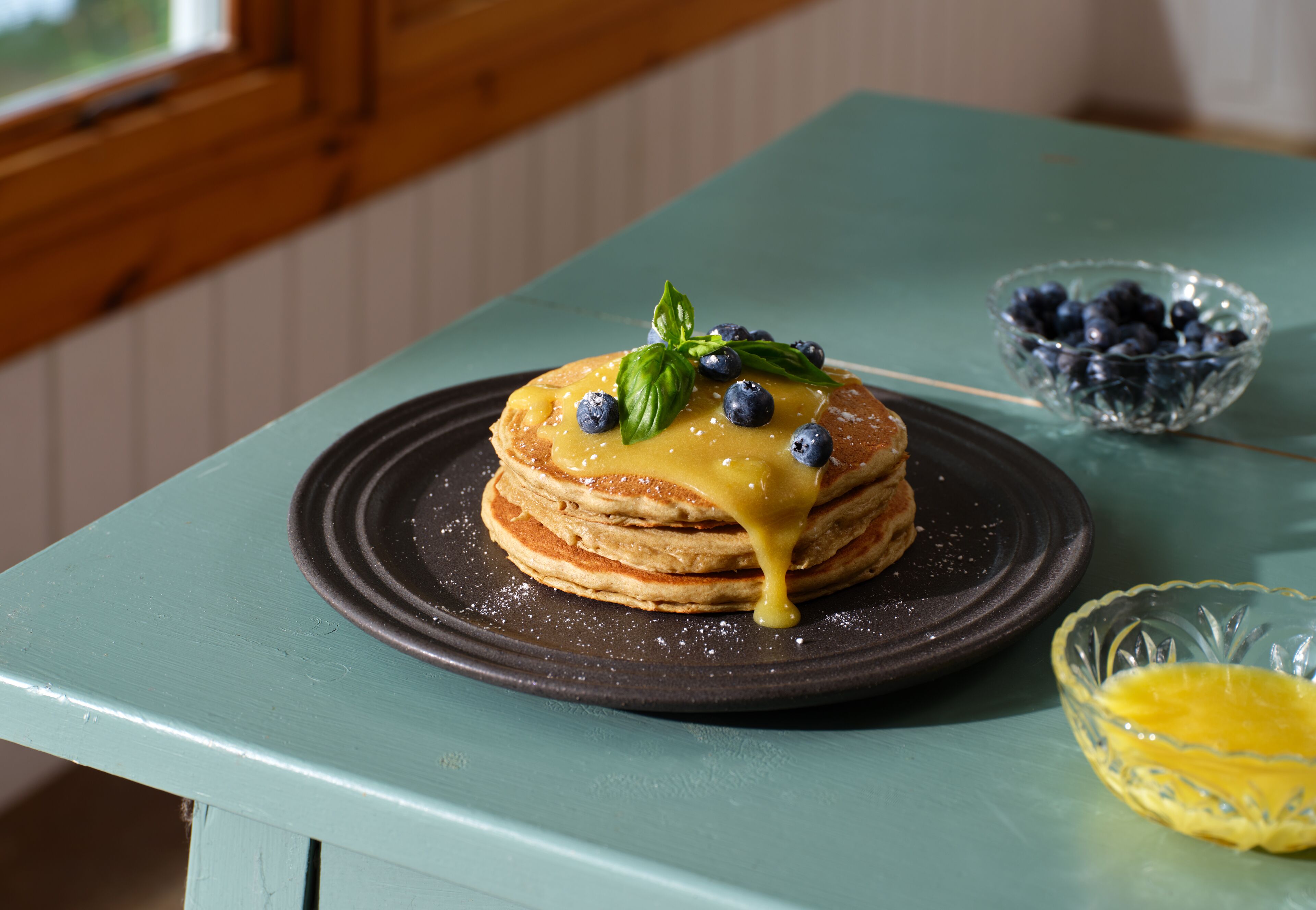 A stack of pancakes on a plate with blueberries and a basil leaf on top, on a teal table.