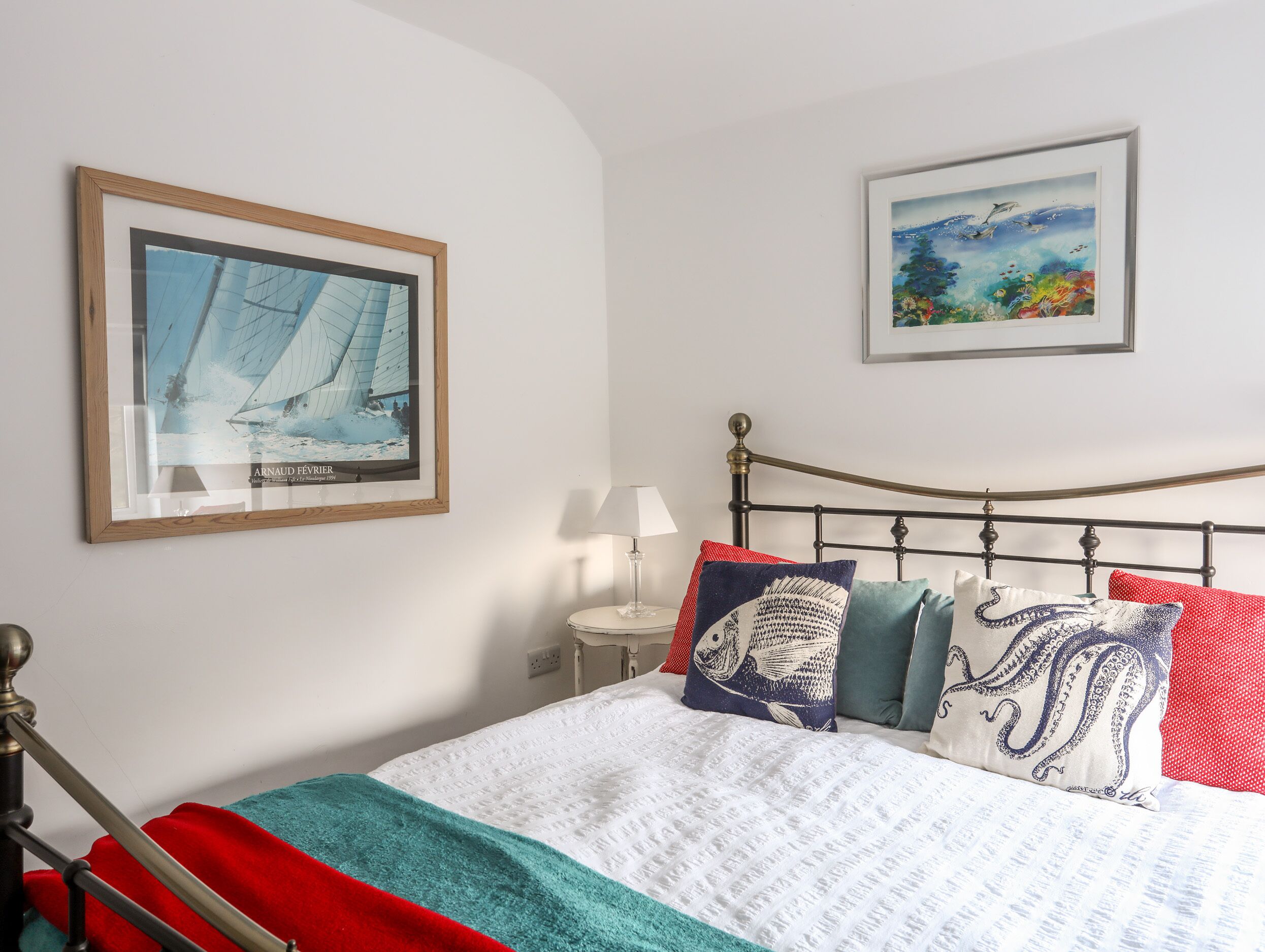 A neatly made bed with a white quilt and colorful pillows, a framed painting of a sailboat, and another painting of a seascape.