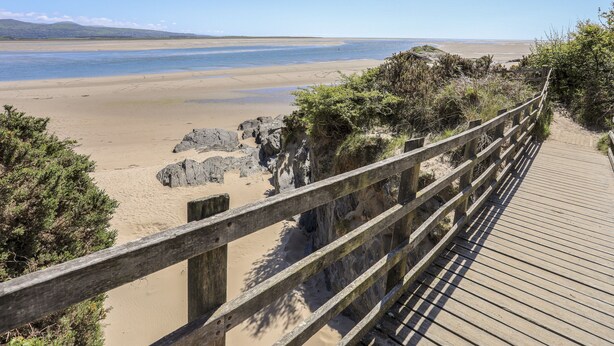 Une promenade en bois qui mène à une plage, avec un ciel dégagé et des montagnes à l’horizon.