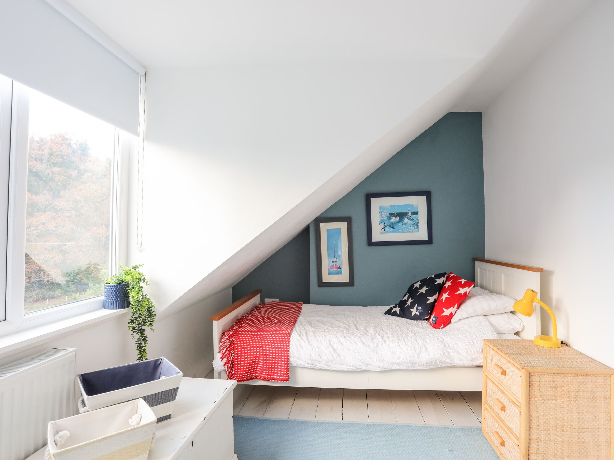 Una camera da letto con soffitto spiovente, un letto con coperta rossa e bianca, una piccola scrivania con cassetti, una lampada da comodino e due quadri appesi al muro.