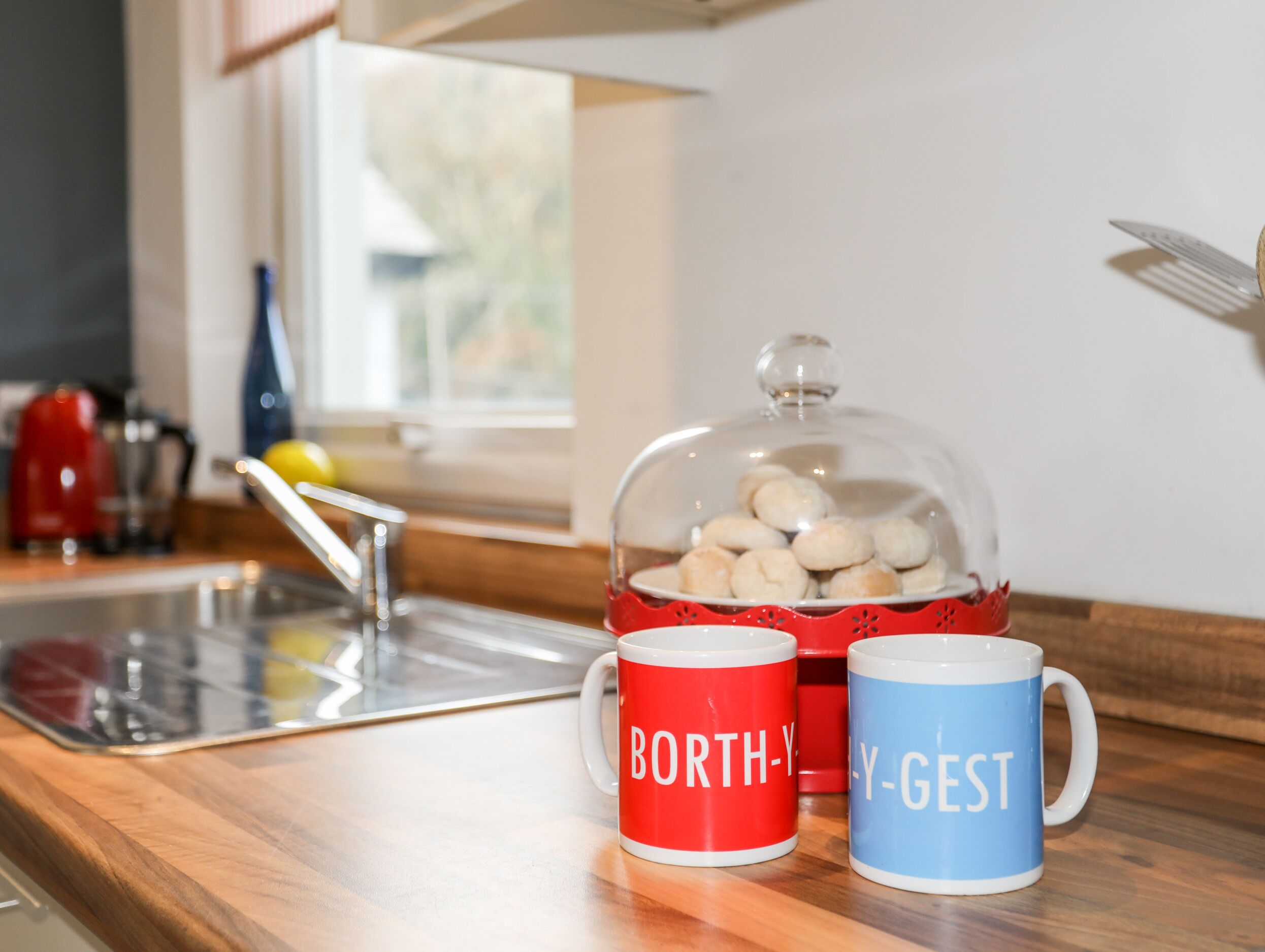 A kitchen counter with two mugs, one red and one blue, and a glass dome covering a plate of cookies.