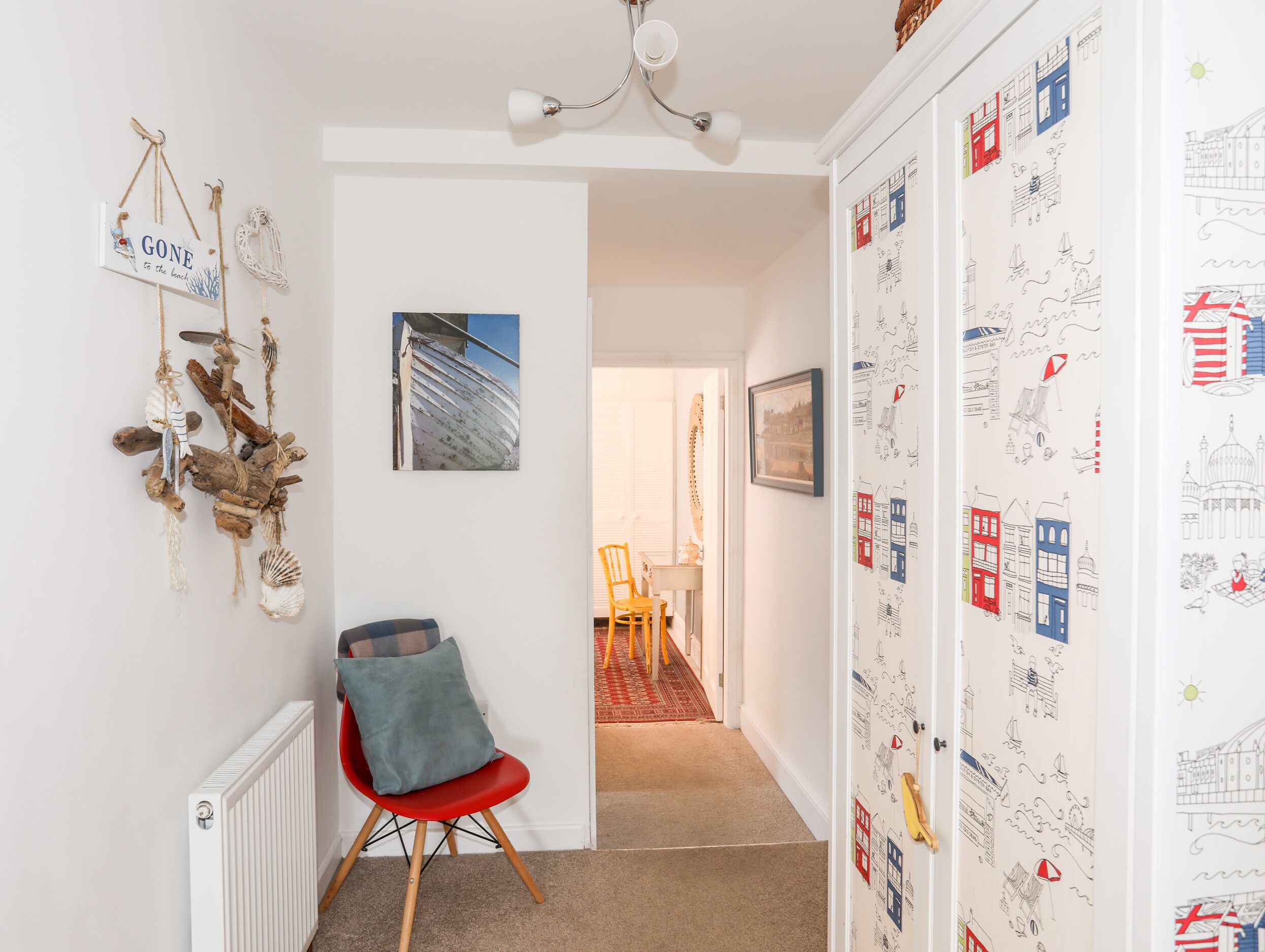 Un couloir avec une chaise rouge, un radiateur blanc, un luminaire au plafond et une porte recouverte d’un papier peint sur le thème du voyage.