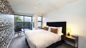 A modern bedroom with a large bed, a nightstand with a lamp, a bench, and a view of the outdoors through large windows. - Lakefront Apartment close to town (Wānaka)