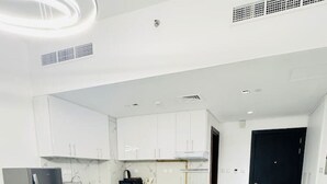 A modern hotel room with a bed, a seating area with a table, a kitchenette, and a dark-colored door.