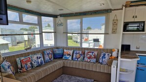 A cozy room with a large window, a wooden bench with nautical-themed cushions, a kitchen area with a microwave, and a small table with a toy duck. - Driftwood Cabin Dog Friendly - Anderby Creek (Skegness)