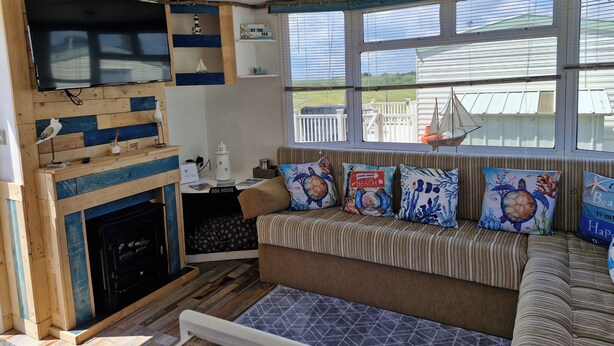 A cozy living room with a fireplace, a television mounted on a wooden wall, a comfortable sofa with beach-themed cushions, and large windows offering a view of the outside. - Driftwood Cabin Dog Friendly - Anderby Creek (Skegness)
