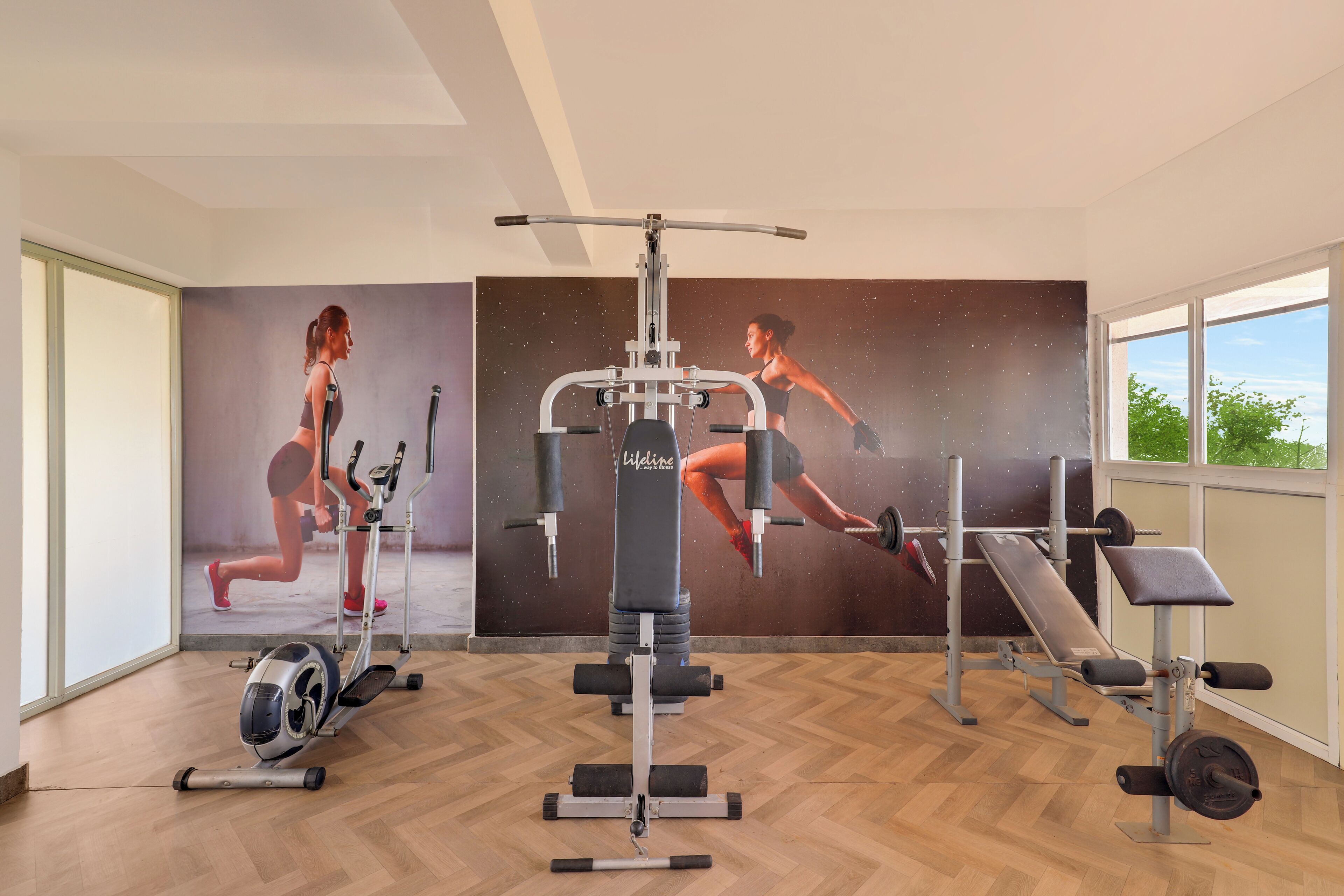 Une salle de sport équipée de matériel d’exercice, un mur orné d’affiches sur le thème du fitness et de grandes fenêtres.
