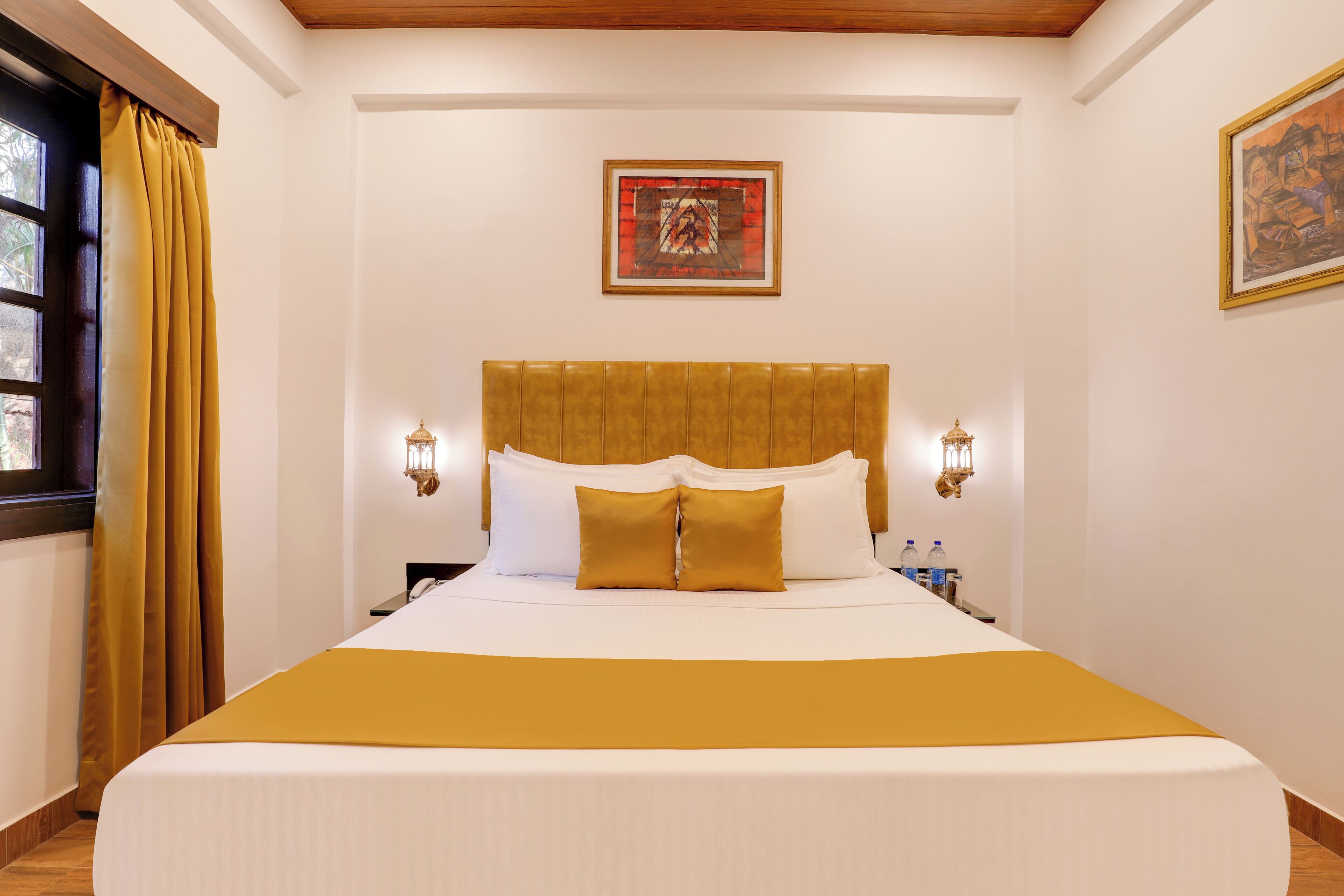 Habitación de hotel con una cama grande, cabecera, dos lámparas de pared, un cuadro enmarcado y una ventana con cortinas.