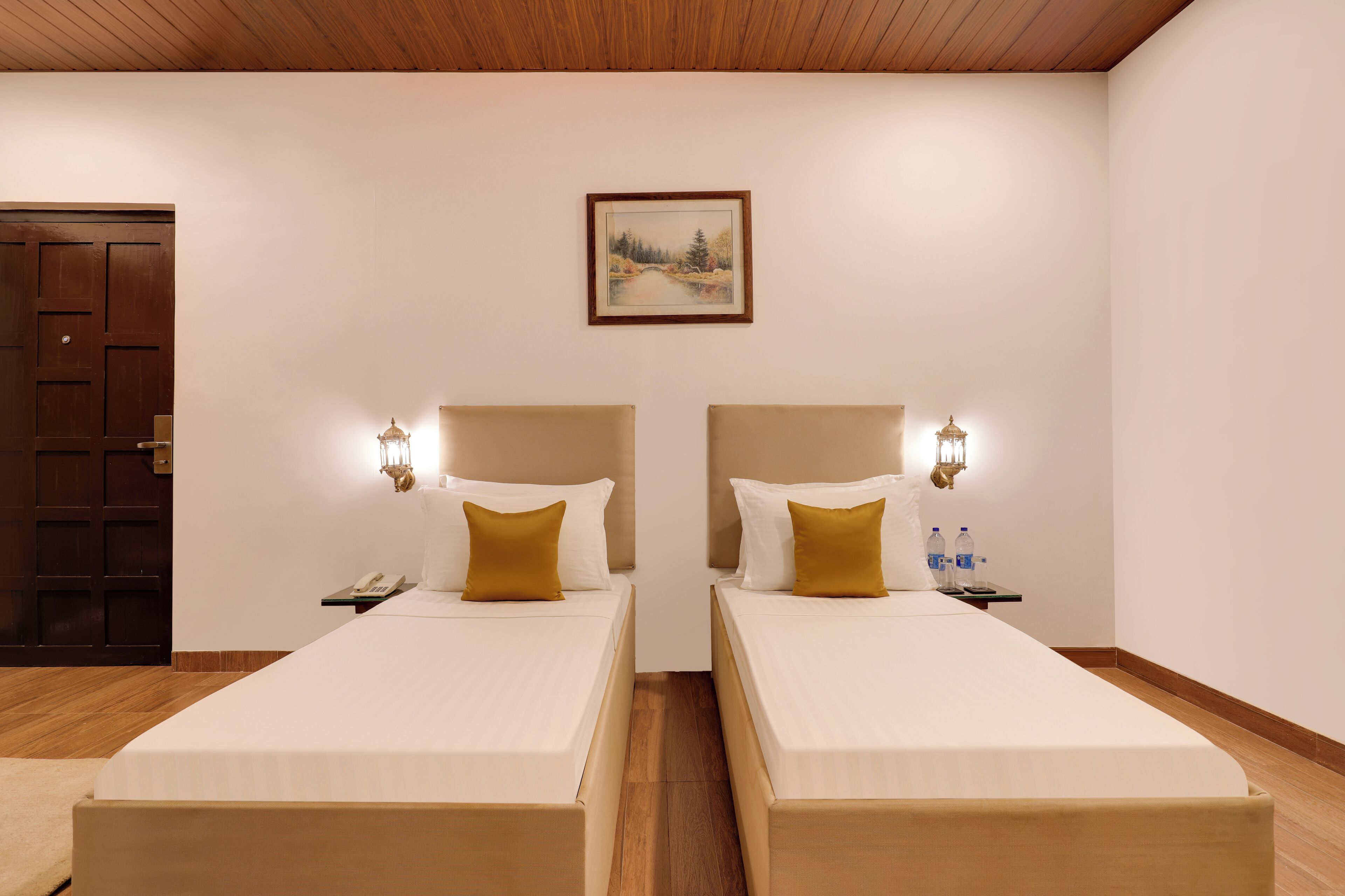 Een hotelkamer met twee enkele bedden, houten hoofdborden, nachtkastjes met lampen, een schilderij aan de muur en een houten deur.