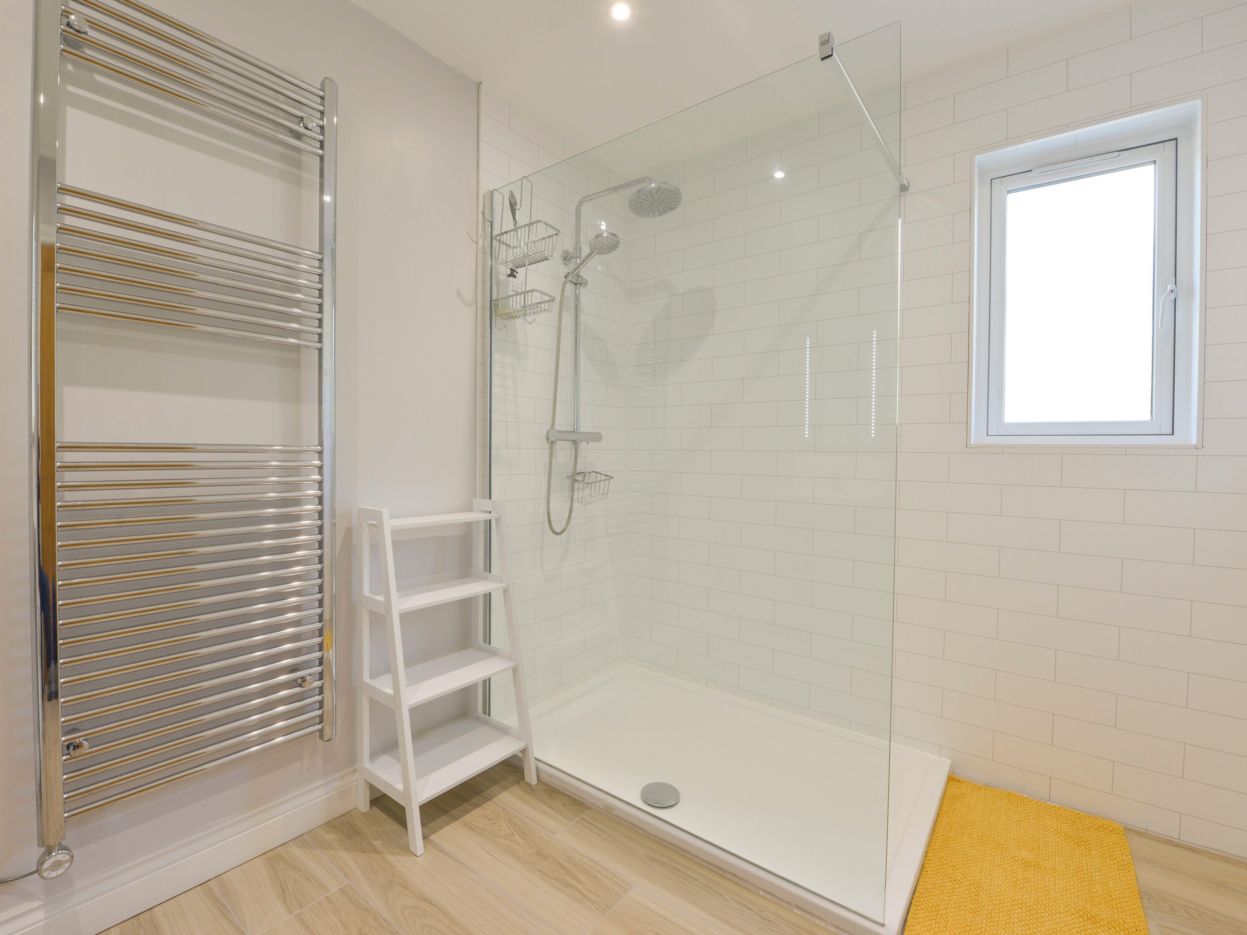 Un bagno moderno con doccia vetrata, parete rivestita in piastrelle bianche, pavimento in legno e una piccola finestra.