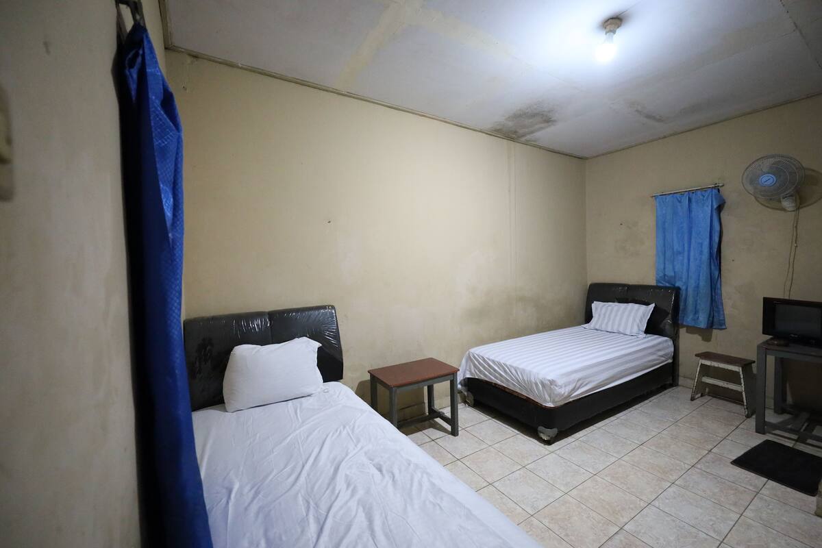 A small, simple room with two beds, a small table, a fan, and a television.
