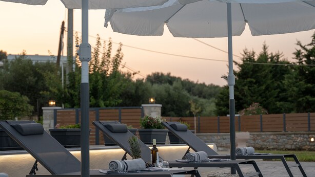 A poolside area with lounge chairs and umbrellas, a stone wall, and a wooden fence in the background. - Villa Nontas Ntina with Private Pool (Rethymno)