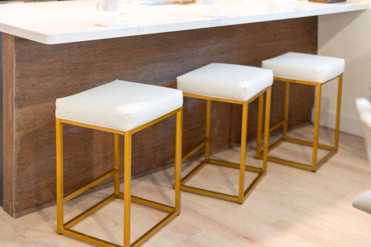 A bar area with three white cushioned stools with gold frames, a white countertop, and a dark wooden backdrop.