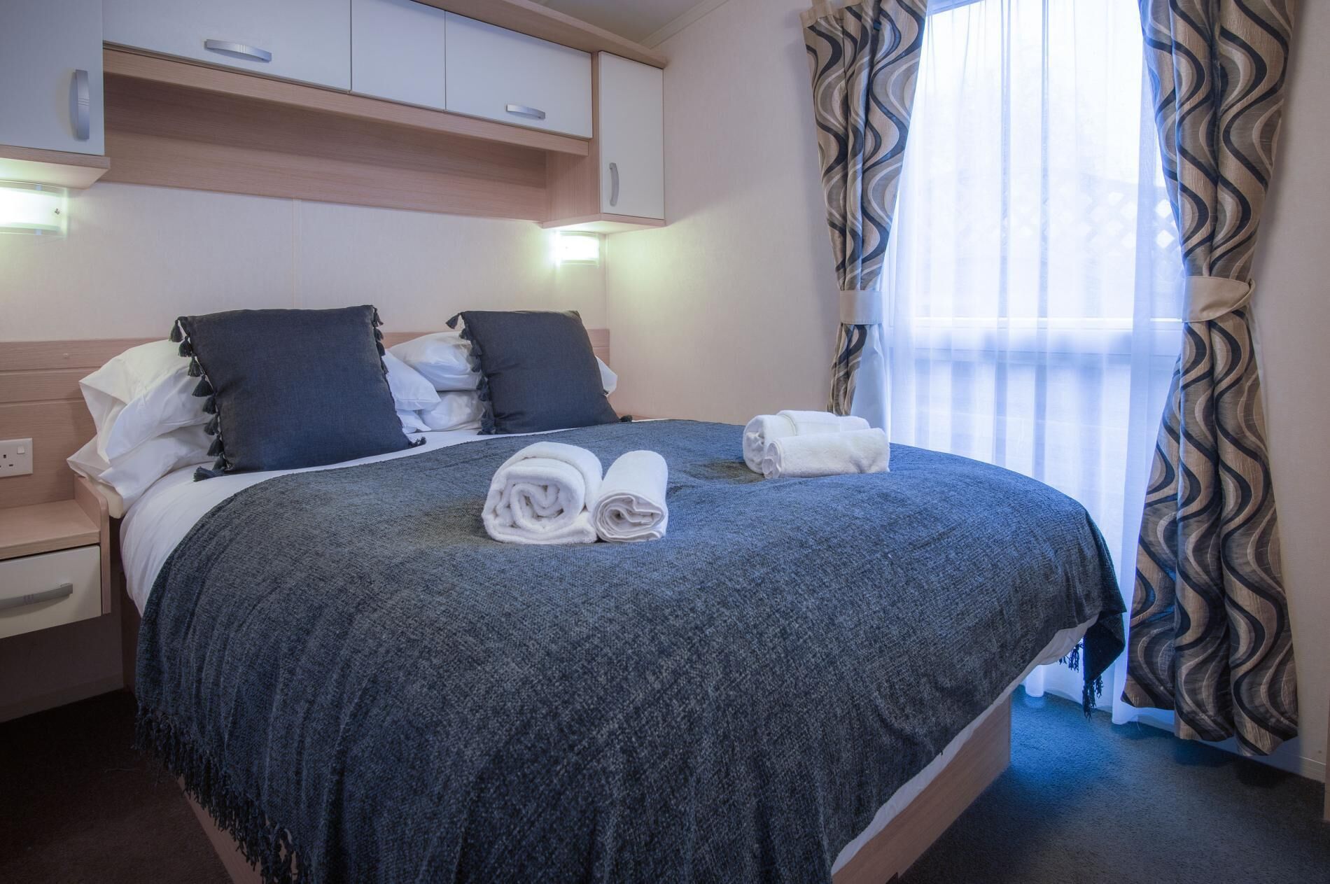 A neatly made bed with a dark blue blanket and pillows, a wooden nightstand, and a window with patterned curtains.