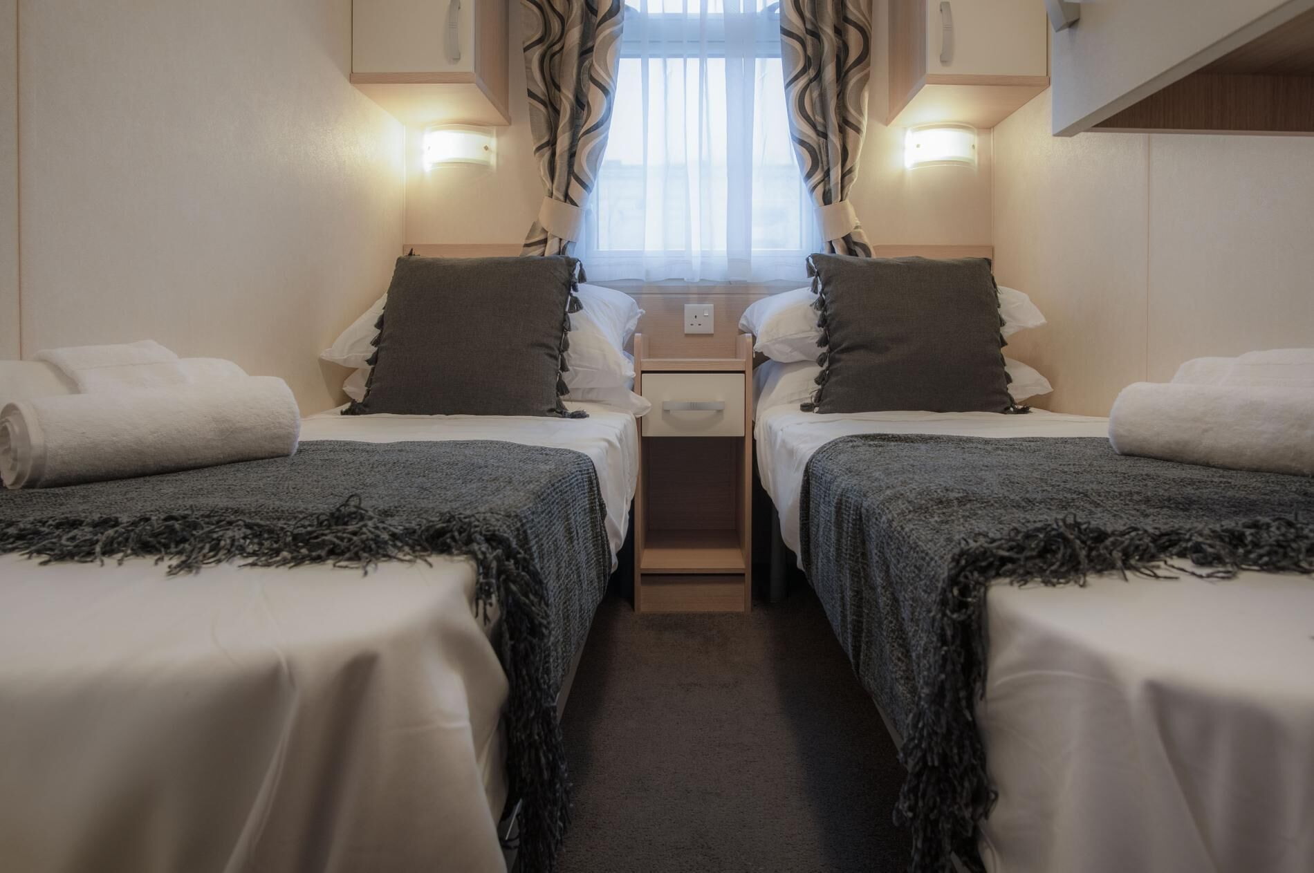 A compact room with two single beds, a small bedside table, a power outlet, and a window with curtains.