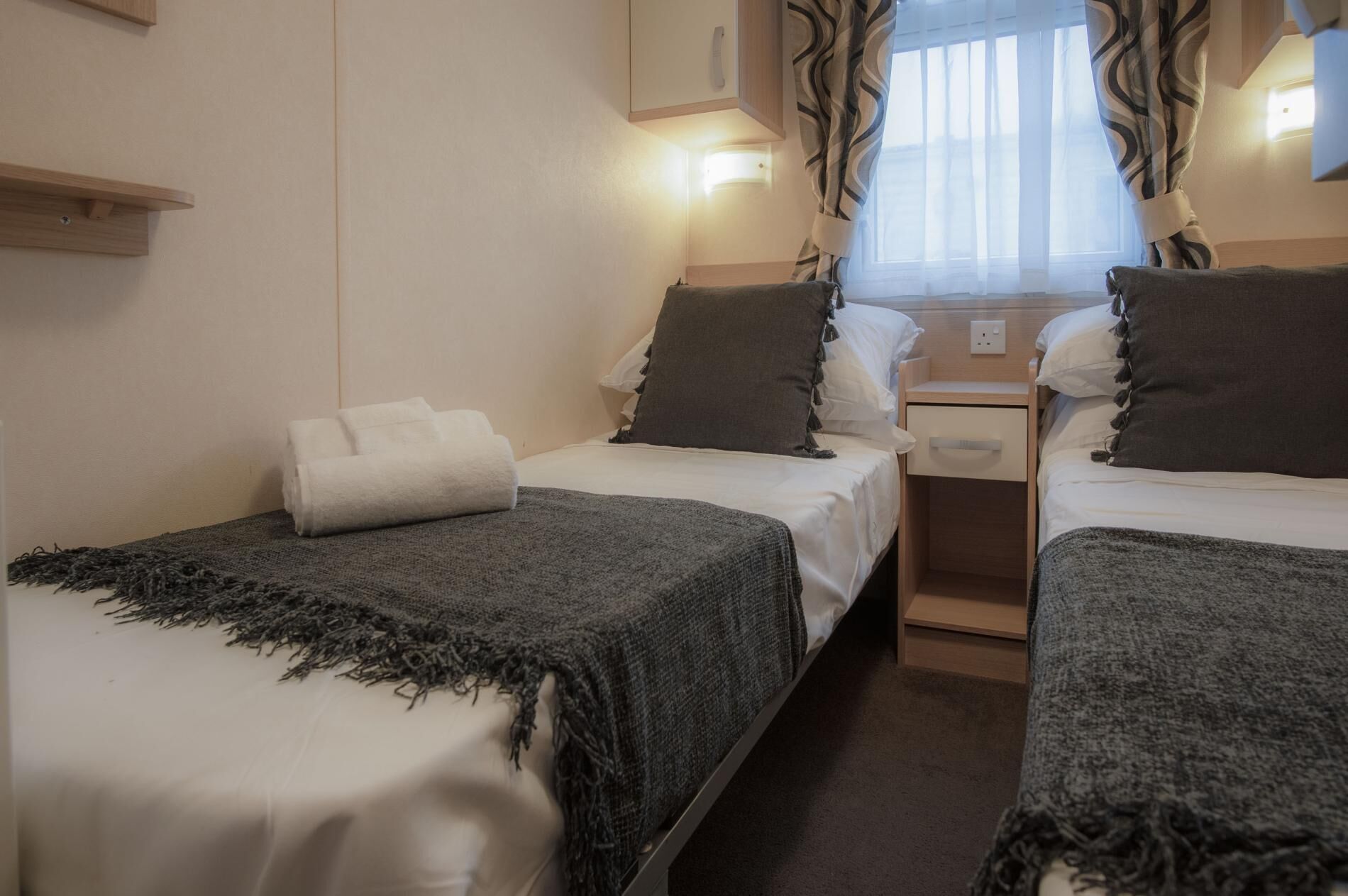 Ein Hotelzimmer mit zwei Betten, jeweils mit grauer Decke und Kissen, einem kleinen Nachttisch mit Lampe und einem Fenster mit Vorhängen.