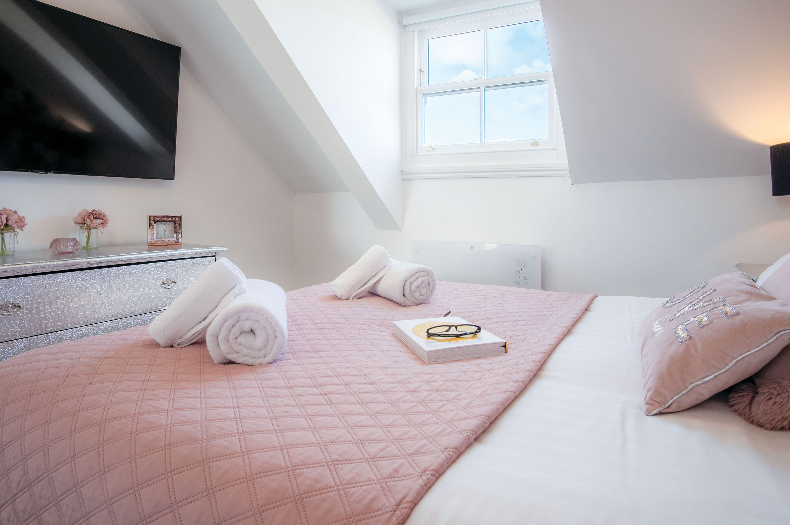 Una cama bien hecha con ropa de cama rosa, un libro y unos lentes encima. Hay toallas enrolladas sobre la cama, un tocador con un espejo y un televisor de pantalla plana montado en la pared.