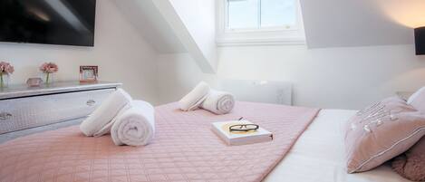 Una cama bien hecha con ropa de cama rosa, un libro y unos lentes encima. Hay toallas enrolladas sobre la cama, un tocador con un espejo y un televisor de pantalla plana montado en la pared.