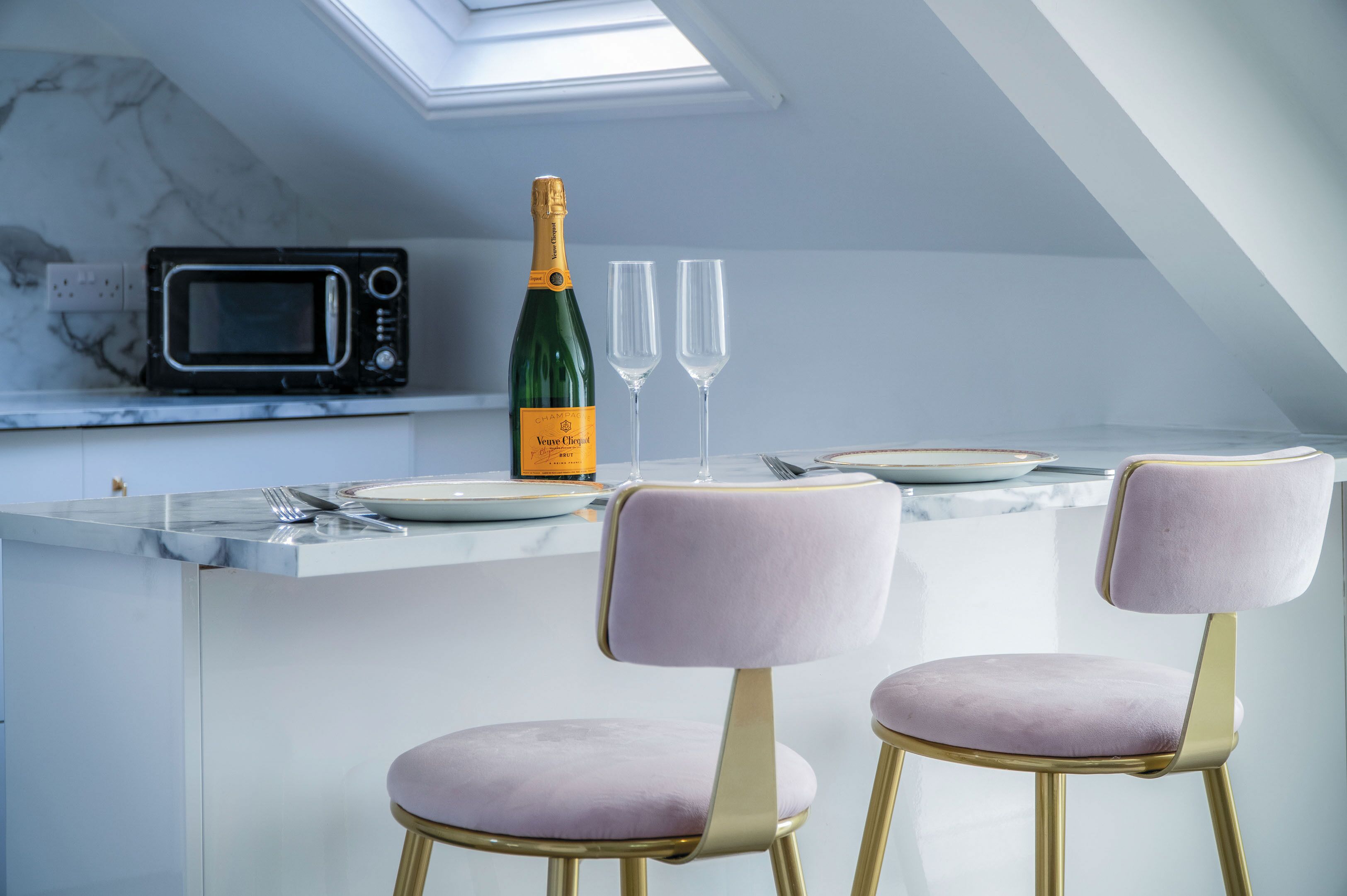 Une cuisine équipée d’un four à micro-ondes, une bouteille de champagne et deux verres posés sur le comptoir. On y trouve deux chaises dotées d’assises rembourrées et de pieds dorés.