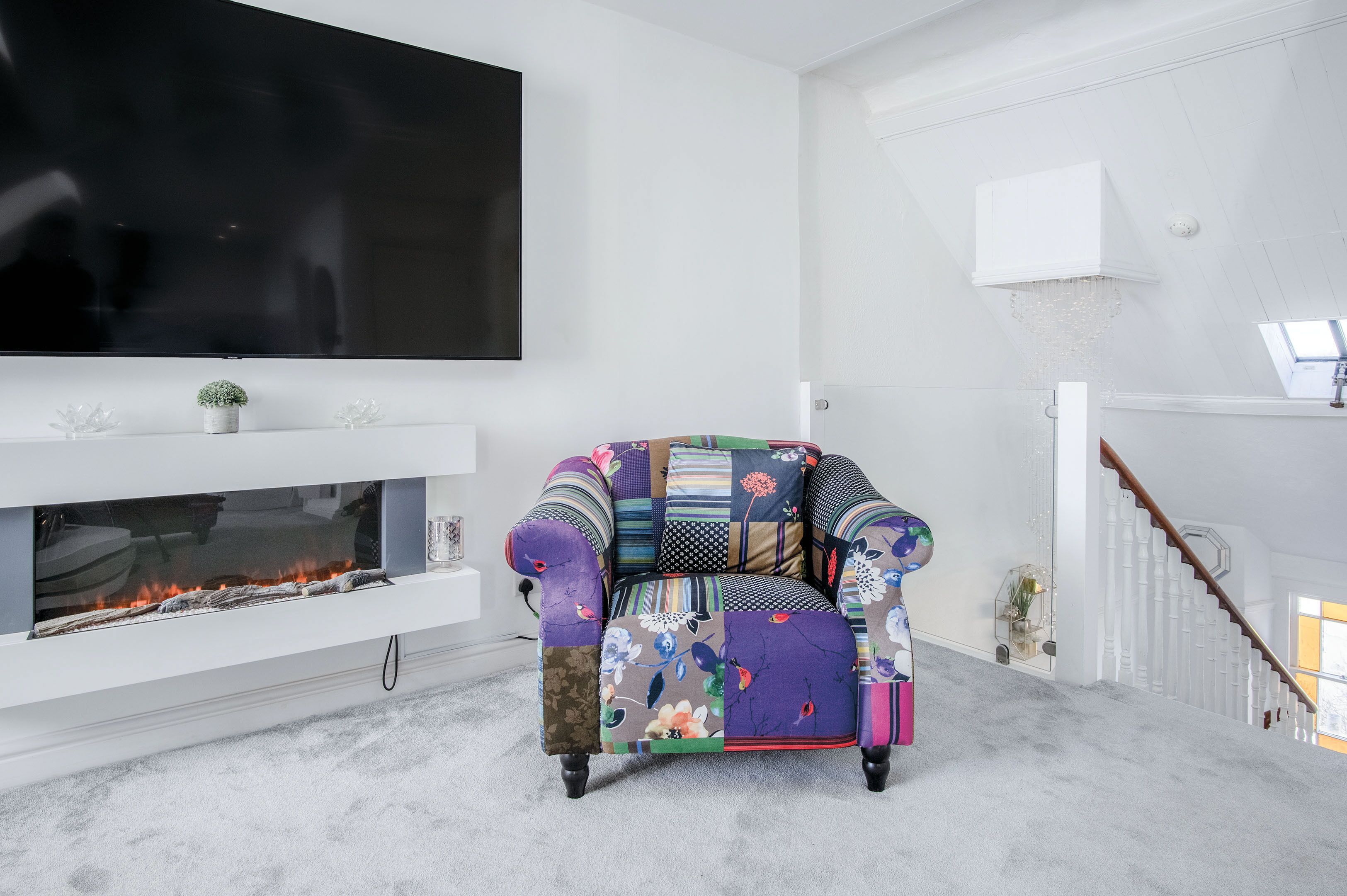 Een woonkamer met een grote flatscreen televisie aan de muur, een modern vormgegeven open haard en een kleurrijke fauteuil met een lappendeken.