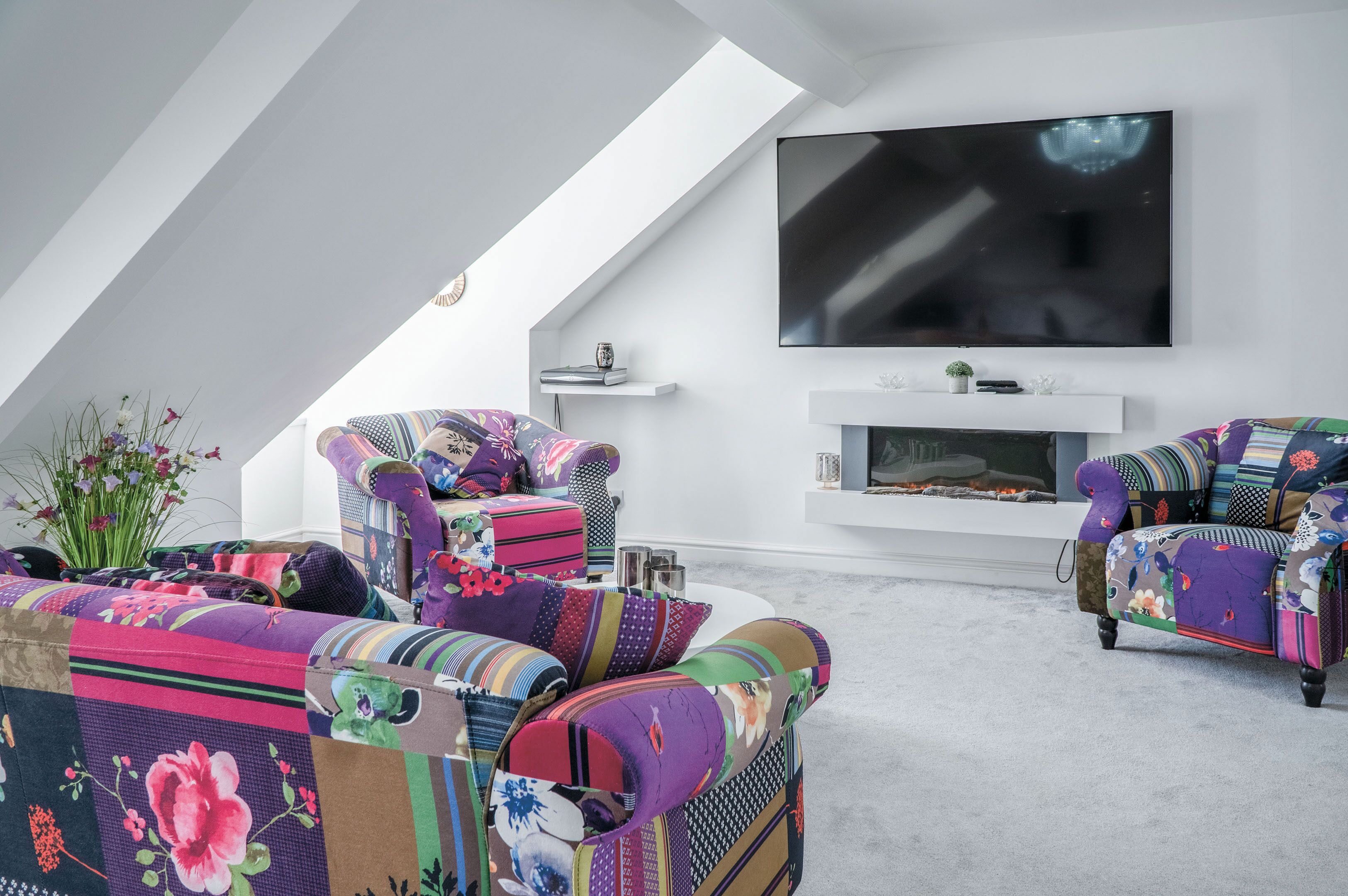 A modern living room with a large flat-screen TV mounted on the wall, a fireplace, and colorful, patterned sofas.
