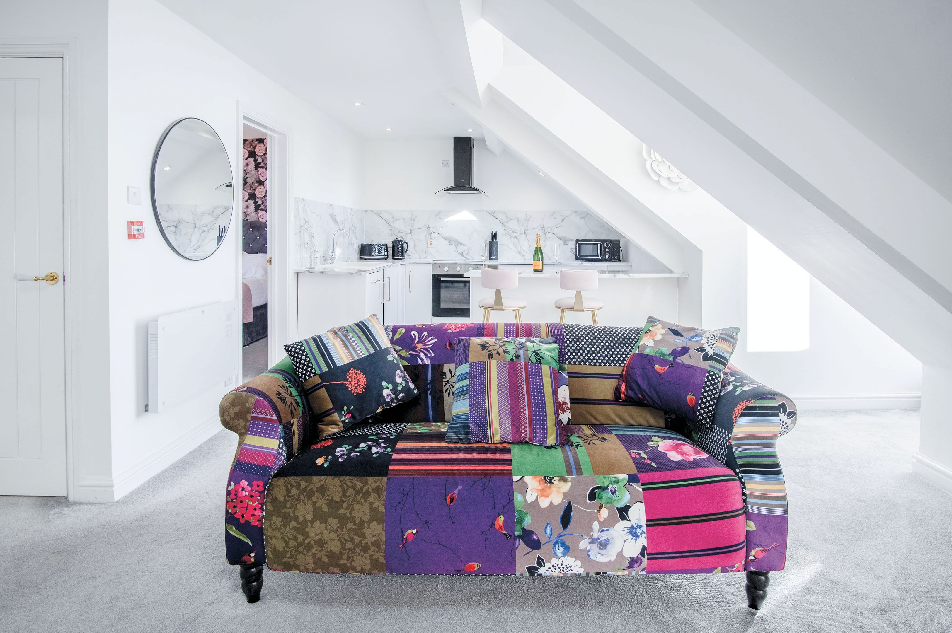 Ein gemütliches Wohnzimmer mit einem farbenfrohen Patchwork-Sofa, einer weißen Tür und einer Küchenzeile mit Mikrowelle und Backofen.