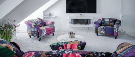 Ein modernes Wohnzimmer mit hohen Decken, zwei gemusterten Sesseln, einem Sofa, einem Flachbildfernseher und einem Kamin.