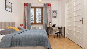 A bedroom with a bed, a desk, and a chair. There is a window with curtains, a radiator, and a picture on the wall.