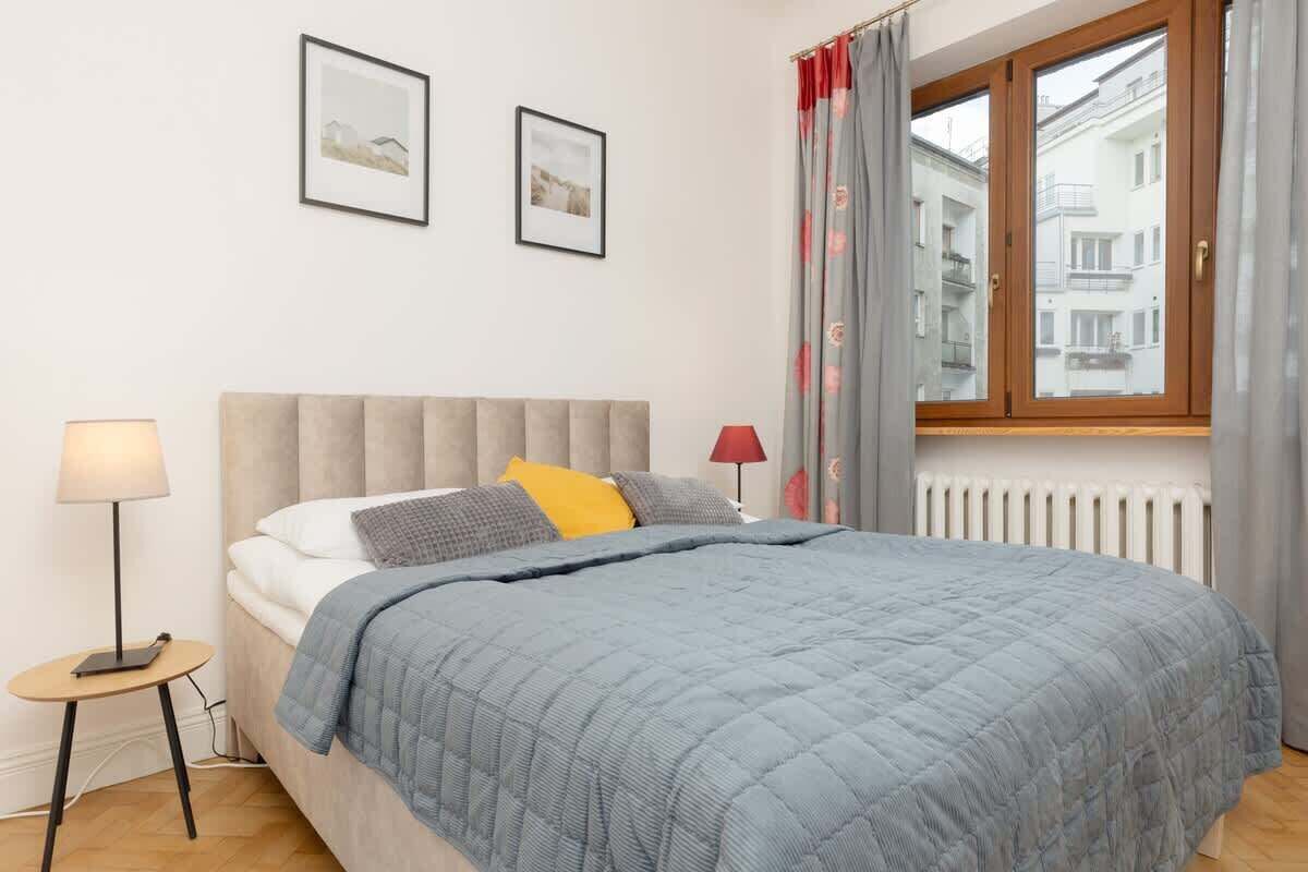 A bedroom with a bed, a bedside table, a lamp, a window with curtains, and two framed pictures on the wall.