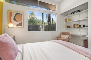 A bedroom with a bed, a chair, a desk, a bookshelf, and a window with a view of trees.