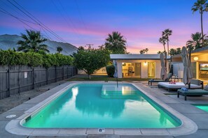 Una piscina rectangular con un indicador de profundidad de 2.4 metros, rodeada de sillas de descanso y sombrillas, con una cerca de madera y un edificio al fondo.
