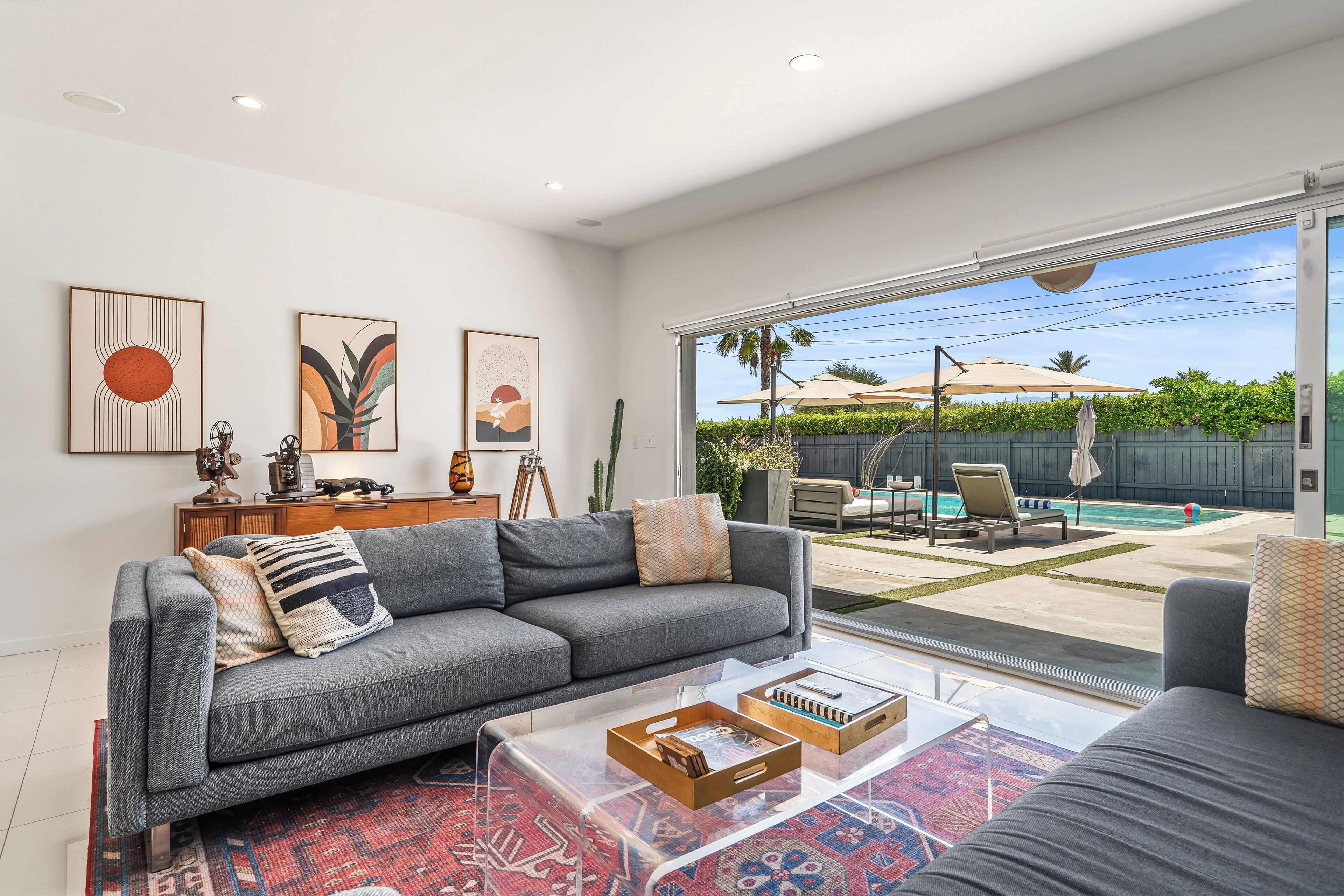 Ein modernes Wohnzimmer mit einem grauen Sofa, einem Couchtisch mit Büchern und einem großen Fenster mit Blick auf einen Garten mit Pool und Gartenmöbeln.