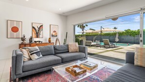 A modern living room with a grey sofa, a coffee table with books, and a large window overlooking a backyard with a pool and outdoor furniture.