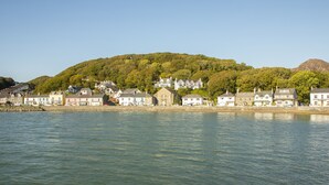 A coastal town with houses along the waterfront, a hill with trees, and a clear sky. - Gweldon - Anchor Deck (Porthmadog)