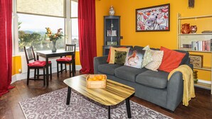 A living room with a sofa, a coffee table, a bookshelf, and a window with curtains. - Gweldon - Anchor Deck (Porthmadog)